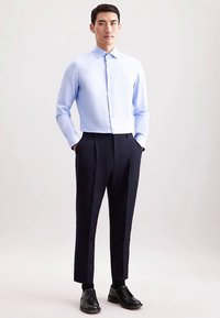 Camicia azzurro chiaro con bottoni e colletto, abbinata a pantaloni sartoriali blu scuro e scarpe eleganti nere. Tessuto liscio, design aderente.