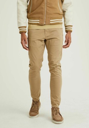Mężczyzna noszący beżowe spodnie typu slim fit, brązowe zamszowe buty sznurowane oraz brązowo-kremową kurtkę varsity, stoi na jednolitym tle.