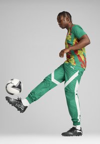 Zaļas sporta bikses ar baltām akcents, kombinētas ar daudzkrāsainu ziedu dizaina krekli. Melni futbola zābaki ar fakturētu virsmu.