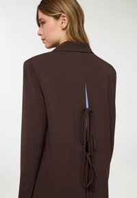 Blazer marrone dal design sartoriale, con spacco posteriore chiuso da anelli di tessuto. Tessuto liscio e maniche lunghe. Interno blu visibile.