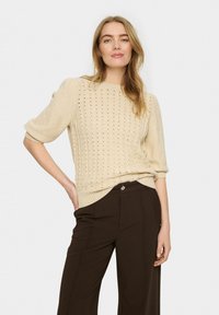 Bežs adīts džemperis ar īsām, pūkainām piedurknēm un atklātām šuvēm, apvienots ar brūnām biksēm ar redzamu pogu dekoru.