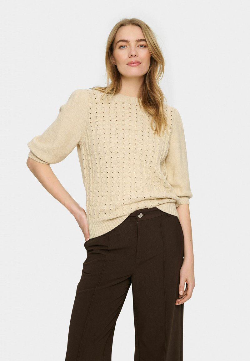 Bežs adīts džemperis ar īsām, pūkainām piedurknēm un atklātām šuvēm, apvienots ar brūnām biksēm ar redzamu pogu dekoru.