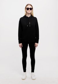 Črna teksturirana kapuca z vrvicami, črne legice in bele športne copate. Model, ki nosi sončna očala, stoji na svetli podlagi.