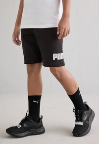 Μαύρες βαμβακερές σορτς με άσπρο λογότυπο "PUMA", συνδυασμένες με μαύρα αθλητικά παπούτσια και μαύρες κάλτσες μέχρι τον αστράγαλο που φέρουν άσπρο λογότυπο.