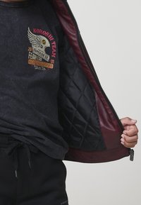 Persona che apre giacca in pelle color bordeaux per rivelare il rivestimento trapuntato nero e una maglietta grigia scura con grafica 'Koroshi Team'.