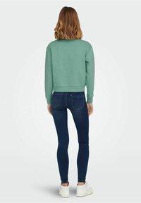 Žena s vlnitými vlasy do délky ramen, oblečená v zelené mikině, tmavých skinny džínech a bílých teniskách, stojící zády k objektivu na jednobarevném pozadí.