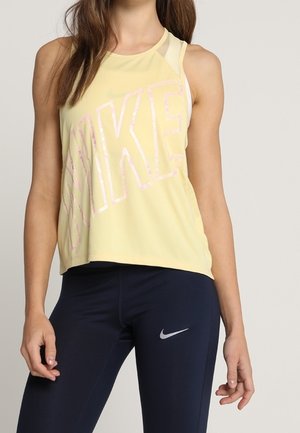 Vrouw draagt een gele mouwloze Nike-top en zwarte Nike-legging, staat met ontspannen armen langs het lichaam tegen een effen achtergrond.