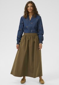 Kvinde med krøllet hår iført en blå langærmet bluse med sommerfuglemønster, olivengrøn maxiskørt og brune slip-on sko, stående.