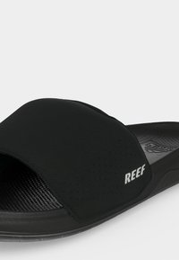 Sandale slide noire à bout ouvert avec semelle texturée et large bride marquée "REEF" sur le côté.
