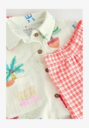 Camicetta a maniche corte color crema con stampa di piante colorate e taschino, abbinata a pantaloni corti a quadri rossi e bianchi.