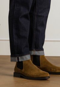 Bruine suède Chelsea boots met zwarte elastische zijpanelen, gedragen met donkere spijkerbroek en opgerolde manchetten, op een houten vloer.