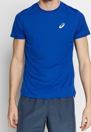 Mężczyzna w niebieskiej sportowej koszulce z krótkim rękawem z logo na piersi i ciemnoszarych szortach, stojący na jasnym jednolitym tle.
