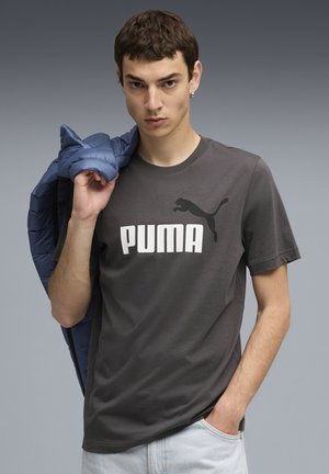 Grå bomulds T-shirt med stort hvidt "PUMA" tekst og sort puma logo. Model holder en blå puffermantel over skulderen. Lys blå denimjeans.