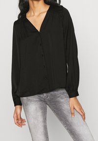 Blusa nera a maniche lunghe con scollo a V, dotata di chiusura a bottoni sul davanti, dettagli plissettati sulle spalle e una texture liscia.