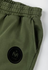 Oliwkowe dresy z elastycznym pasem, z czarną okrągłą łatą z logo oraz miękkim, teksturowanym materiałem. Widoczne kieszenie.