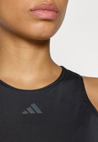 Czarny sportowy top na ramiączkach z wysokim dekoltem, z tonalnym logo w formie trzech pasków i gładką, matową teksturą. Smukły design bez dodatkowych detali.