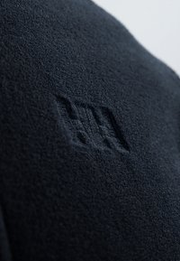 Crni flis materijal s mekom teksturom, s reljefnim logotipom za detalj. Materijal izgleda debelo i toplo.