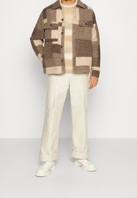 Mand iført brun og beige jakke med geometrisk mønster, beigefarvet stribet sweater, hvide bukser med vide ben og hvide sko, stående mod en hvid baggrund.