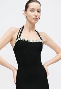 Μαύρο φόρεμα με τιράντες στον λαιμό, κατασκευασμένο από ριμπ ύφασμα, με λαιμόκοψη διακοσμημένη με κρυστάλλους και εφαρμοστή γραμμή. Ομαλή υφή, χωρίς μανίκια.