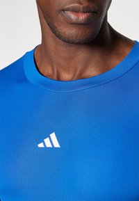 Kék sportpóló sima, könnyű anyagból. Kerek nyakú fazon, az alsó bal sarokban fehér Adidas logóval.