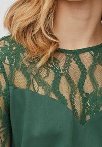 Capelli biondi su un vestito verde con dettagli in pizzo floreale e una rete trasparente sul collo e le spalle.
