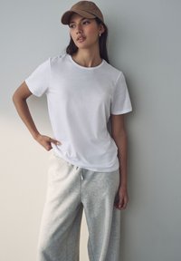 T-shirt bianco a maniche corte abbinato a pantaloni da jogging grigio chiaro. Modello che indossa un cappello beige, in piedi contro un muro neutro. Look casual.