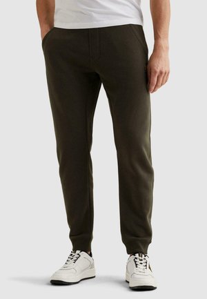 Man draagt donker groene joggerbroek en witte sneakers, staat met handen in zakken op een lichte achtergrond.