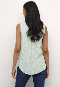 Bluza fără mâneci de culoare verde deschis, cu un model floral, tiv curbat și croi lejer, asortată cu blugi din denim de culoare albastru închis.