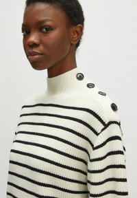 Pull en maille avec un col montant, présentant des rayures horizontales noires et blanches, et des boutons décoratifs noirs sur l'épaule.