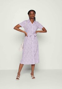 Mulher com cabelo trançado, vestindo um vestido midi floral lilás com mangas curtas, gola, cinto à cintura, sandálias brancas e uma pequena bolsa de ombro branca.