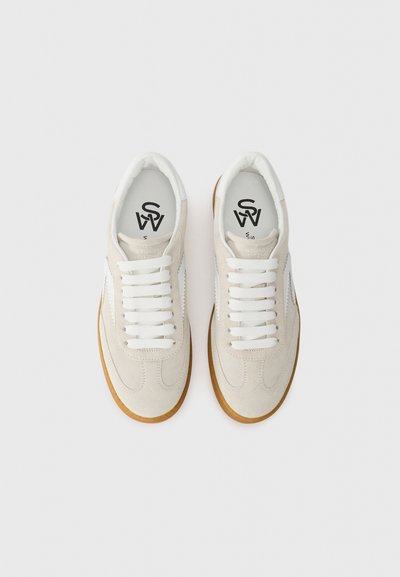 Baskets en suède blanc avec une semelle en gomme, présentant des lacets blancs et un logo sur la languette. Le design comprend une bande latérale texturée.