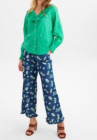 Camicia verde con colletto arricciato; pantaloni a palazzo blu con motivo floreale e orli arricciati; sandali con tacco marroni.