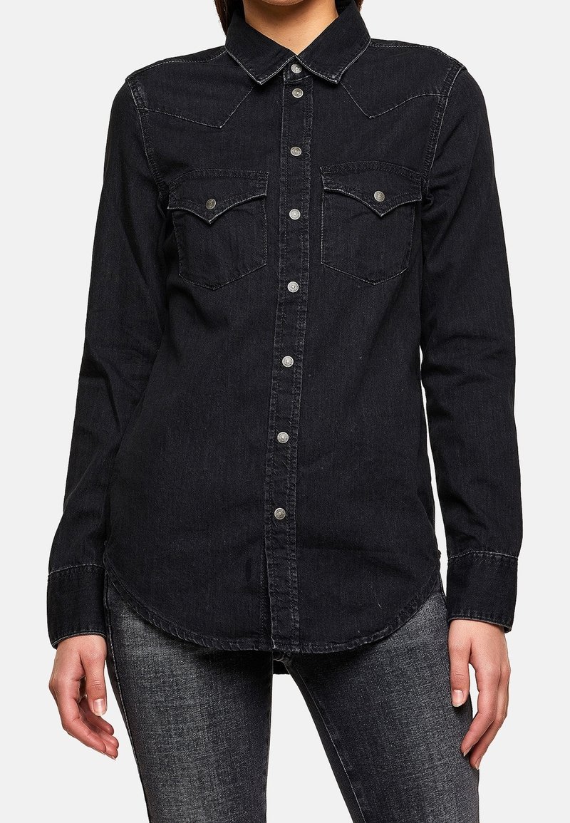 Personne portant une chemise noire en denim à manches longues et boutons, avec des poches poitrine, et un jean gris-noir, montrée des épaules aux cuisses.