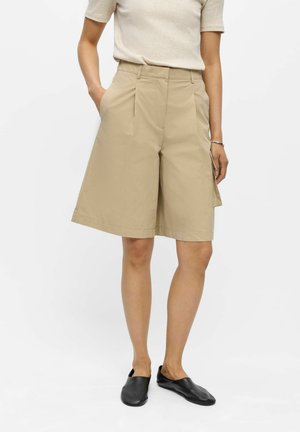 Personne portant un short beige à jambes larges, un haut beige clair à manches courtes et des chaussures noires sans lacets, debout avec une main dans la poche.