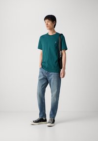Teal cotton t-shirt with a crew neck and small logo on the chest, paired with light blue denim jeans and black sneakers.
