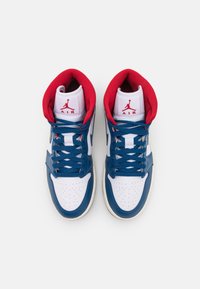 Paire de baskets montantes Air Jordan blanches et bleu marine avec doublure intérieure rouge et lacets bleus, placées côte à côte sur un fond blanc.
