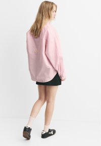 Camicia oversize a righe di colore rosa chiaro, con maniche a polsini, indossata su una gonna nera. Presenta un ricamo sottile sulla schiena. Sneakers nere con dettagli bianchi.