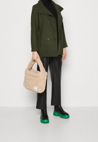 Persona vestida con abrigo verde oscuro, cuello alto negro, pantalones de cuero negros, botas negras con suelas verdes, sosteniendo un bolso de piel beige y esponjoso.