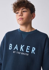 Marineblå sweatshirt i vaffelstrik med rund halsudskæring, med lyseblå "BAKER" broderi og "by TED BAKER" i mindre tekst nedenunder.