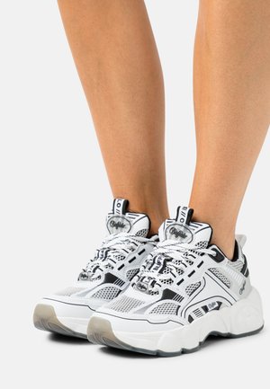 Zapatillas deportivas en blanco y negro que presentan material de malla, acentos texturizados, suela gruesa y un logotipo audaz en la lengüeta y los lados.