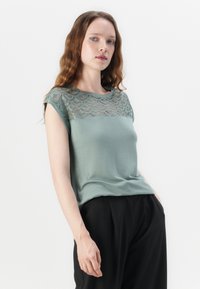 Mintgrønn skjorte med en blonder-overside med en bølget mønster på skuldrene, korte ermer og en myk, glatt tekstur.