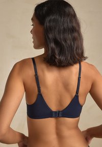 Tamsiai mėlynas bevielis liemenėlė su plonais reguliuojamais dirželiais, lygiu audiniu ir kablys-akis užsegimu, sujungtu maža kaspino detale nugaroje.