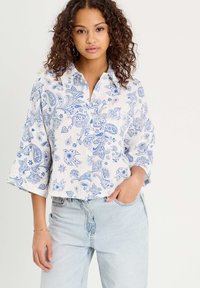 Chemise blanche à boutons avec des motifs paisley bleus, col, et manches trois-quarts, associée à un jean bleu clair.
