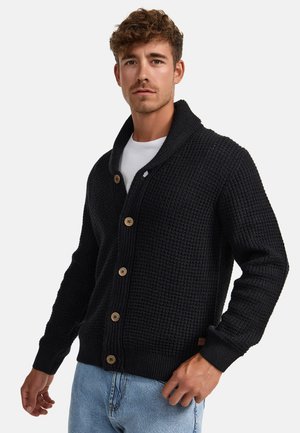 Strikket svart cardigan med sjalkrage, prydet med treknapper, ribbede mansjetter og en teksturert vev, kombinert med en hvit t-skjorte og jeans.