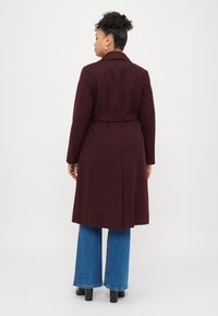 Μπορντό παλτό με ζώνη στη μέση και μακριά μανίκια, σε συνδυασμό με φαρδιά μπλε τζιν και μαύρες μποτάκια με τακούνι. Λείος υφασμάτινος ιστός.