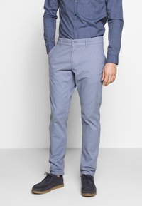 Man som bär ljusblå smala byxor, marinblå mönstrad skjorta med knappar, och mörkblå snörskor, står med händerna i fickorna.