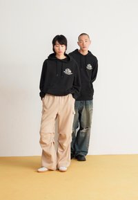 Two models wear hoodies: one in black with white logo, the other in beige and distressed blue jeans. Background is plain white, floor is yellow.