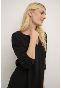 Černý top s dlouhým rukávem, výstřihem do V a ohrnutými manžetami. Měkká tkanina s volným střihem. Mezi výrazné detaily patří knoflík na každé manžetě.