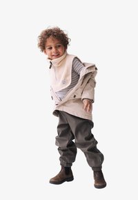 Beige wetterfeste Jacke mit hohem Kragen, über ein grau gestreiftes Hemd geschichtet. Dunkelgrüne Hose mit elastischem Bund und braune Ankle-Boots.