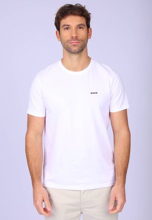 Homme aux cheveux courts et foncés portant un t-shirt blanc uni avec un petit logo noir d'animal sur la poitrine, debout devant un fond clair.